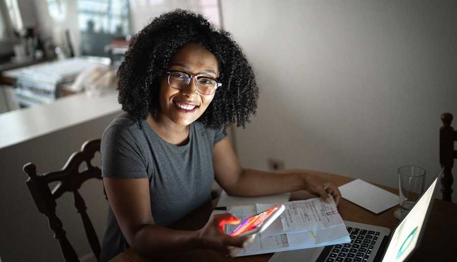 smiling teleworker sits at dining table with laptop, papers and mobile device 