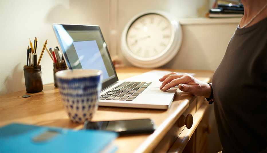 A woman sits at a desk at home with a hot drink and phone and uses a laptop, close up