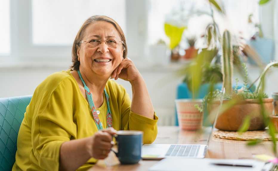 a smiling woman in a yellow sweater holds a blue coffee cup while sitting at a laptop