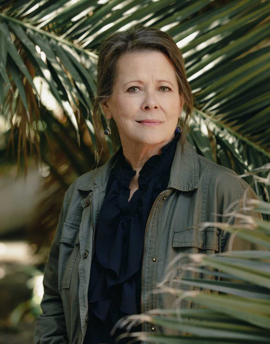 elena medo stands amid fronds while wearing an olive jacket and a black shirt