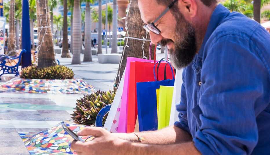 Hombre sentado en una banca con bolsas de compras y viendo su teléfono móvil.