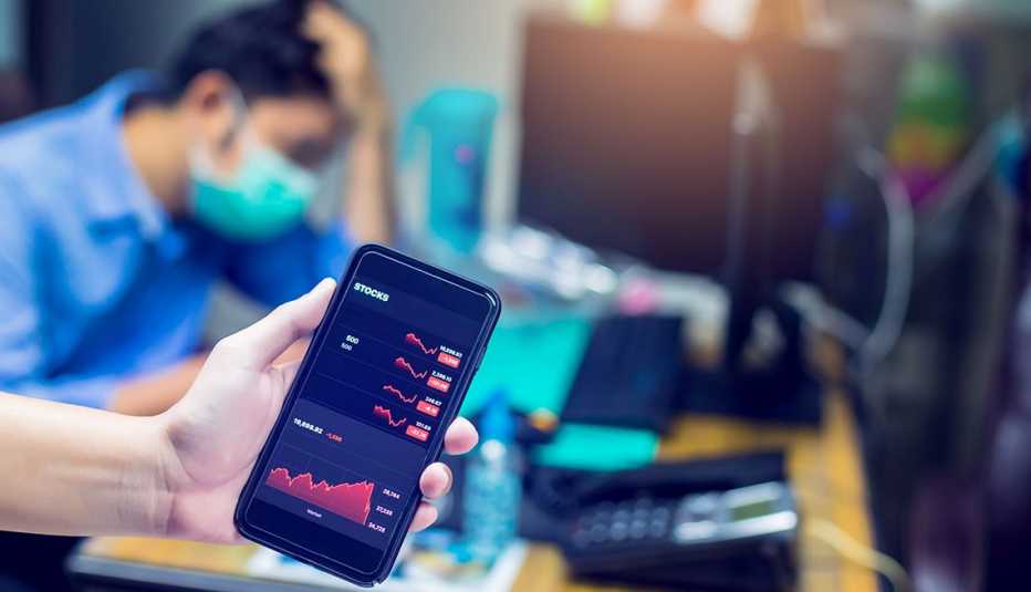 Teléfono móvil con gráficas del mercado de valores y al fondo un inversionista con un tapabocas.