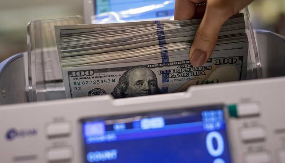 Máquina contando billetes de 100 dólares que son sostenidos por una mano.