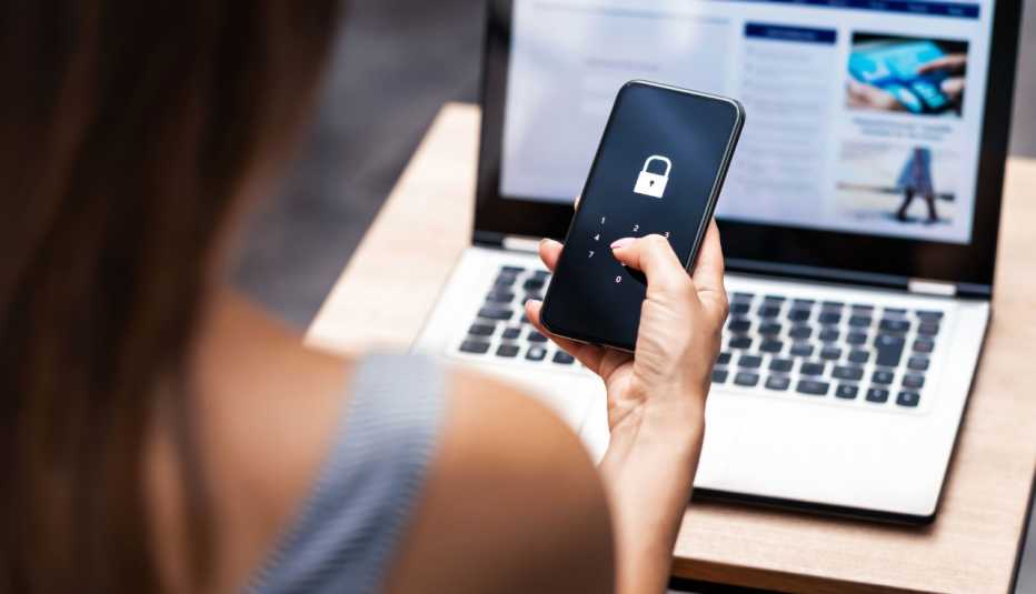 Mujer sostiene un teléfono móvil con un candado en la pantalla frente a una computadora