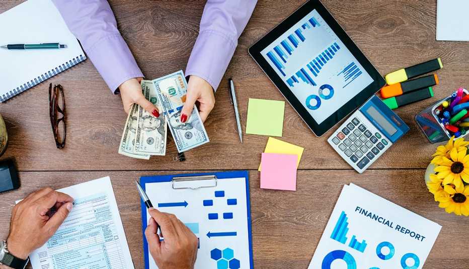 Mesa con las manos de dos personas haciendo cuentas, con calculadoras, formularios y dinero.