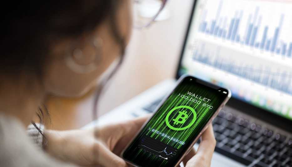 Mujer mirando un símbolo de bitcóin en la pantalla de su teléfono y con una computadora alfondo.