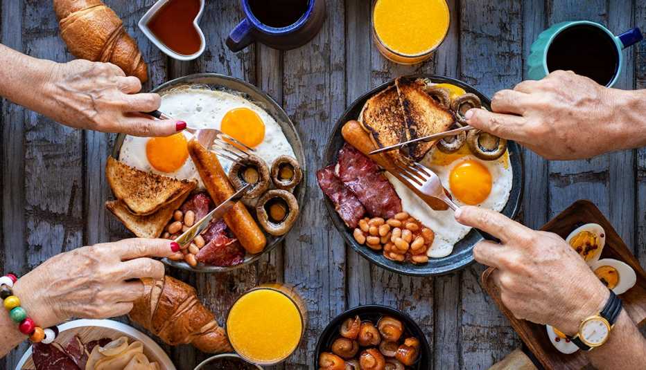 Mesa servida con platos de un desayuno tradicional y manos de personas con cubiertos sobre ellos. Mesa servida con platos de un desayuno tradicional y manos de personas con cubiertos sobre ellos.