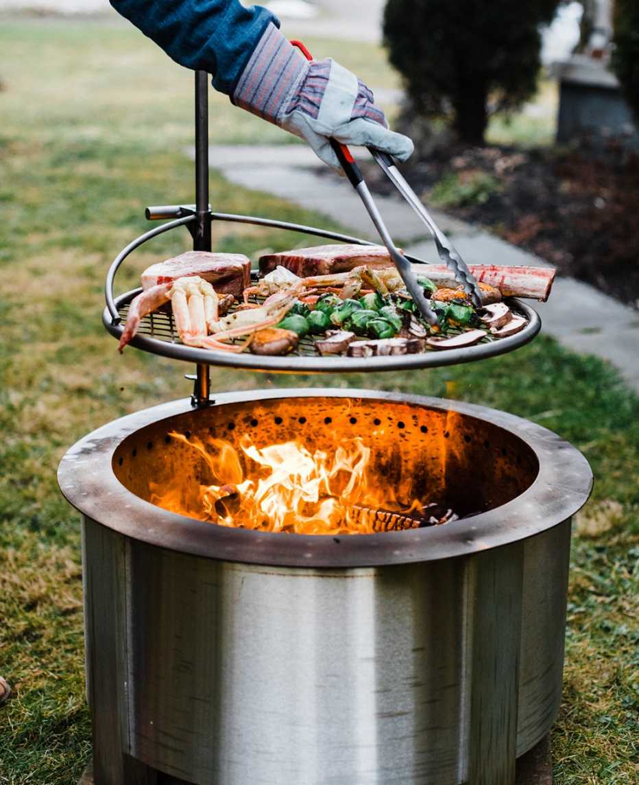 Persona cocinando carne y vegetales encima de un pozo de hoguera Breoo 