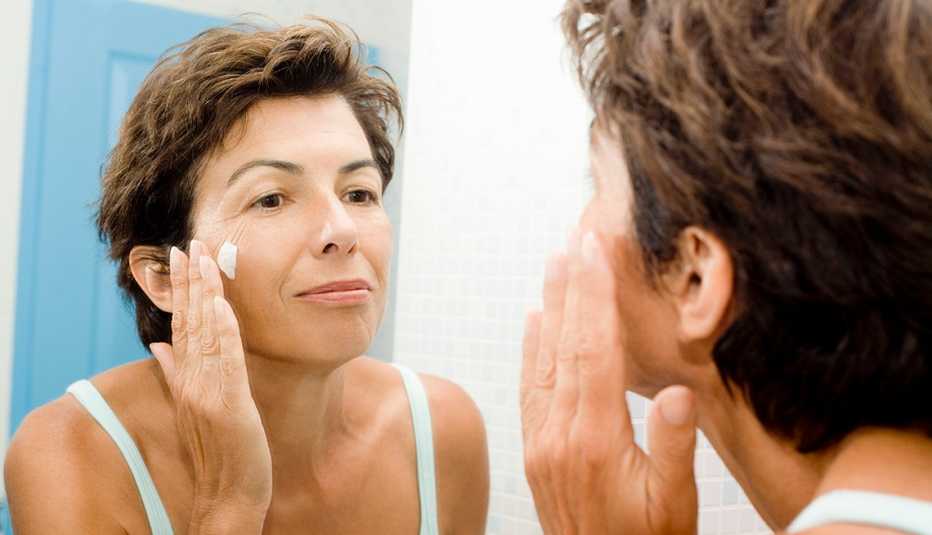 Mujer mirándose en un espejo mientras se aplica una crema en la cara.