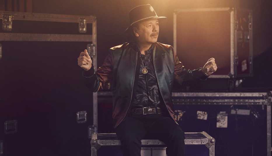 Carlos Santana sentado sobre cajas de equipo musical y tronando sus dedos en el aire.