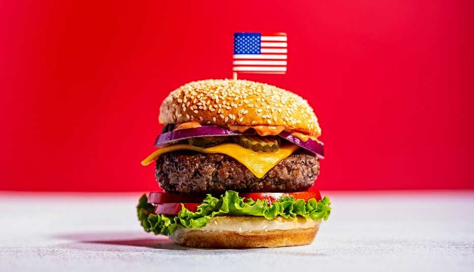 Una hamburguesa con carne, queso y verduras en un pan con semillas de sésamo y con un palillo con la bandera estadounidense clavado en la parte superior.