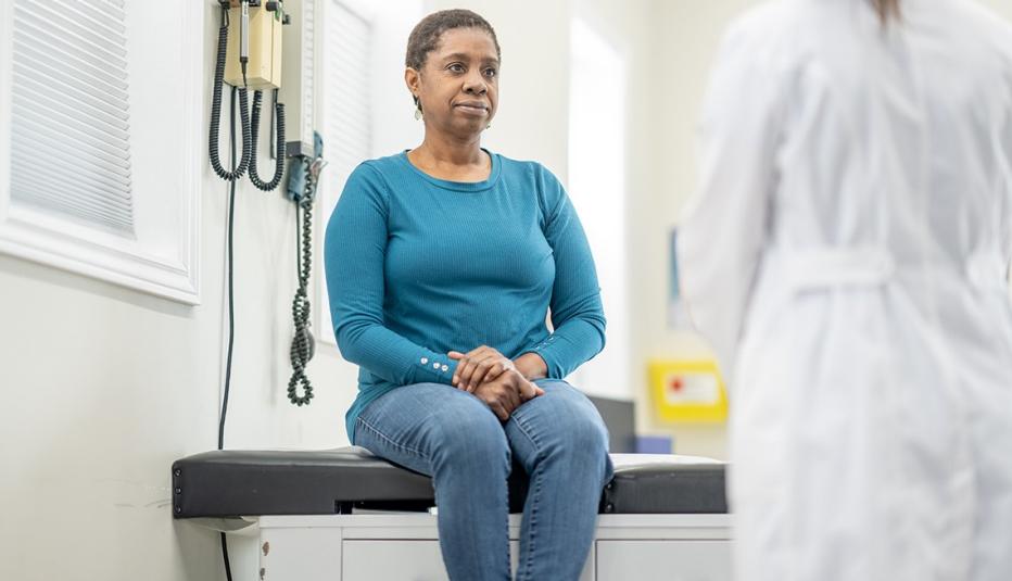 A senior woman of African decent, sits up on an exam table during a routine check-up as she greets her doctor. She is dressed casually and smiling as her female doctor enters the room.