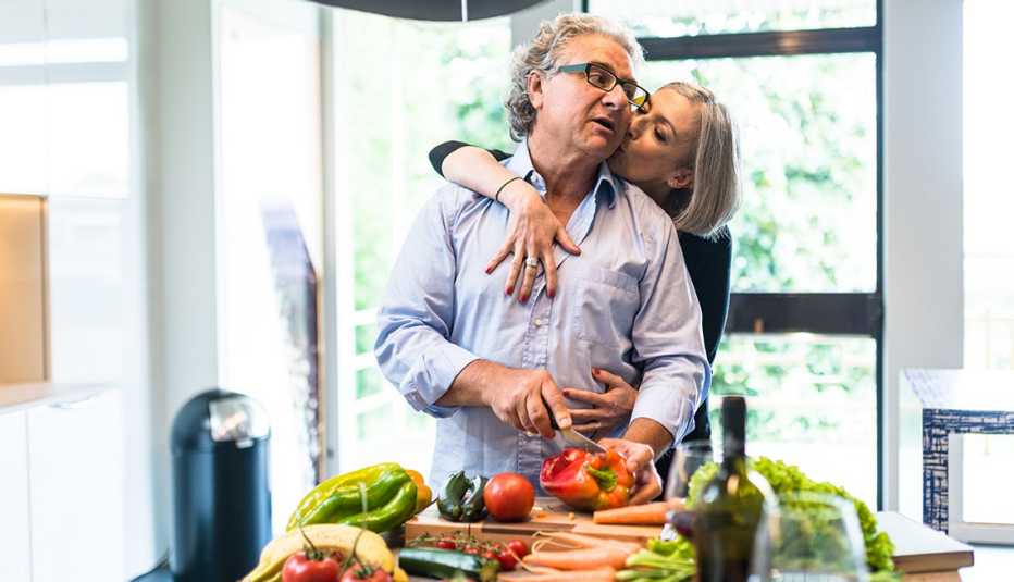 Hombre cocinando en su casa mientras su esposa lo abraza por detrás y le da un beso. Hombre cocinando en su casa mientras su esposa lo abraza por detrás y le da un beso.