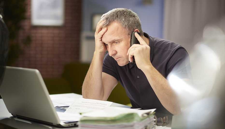Hombre mirando una computadora mientras que con una mano se sostiene la cabeza y con la otra sostiene un teléfono en su oído