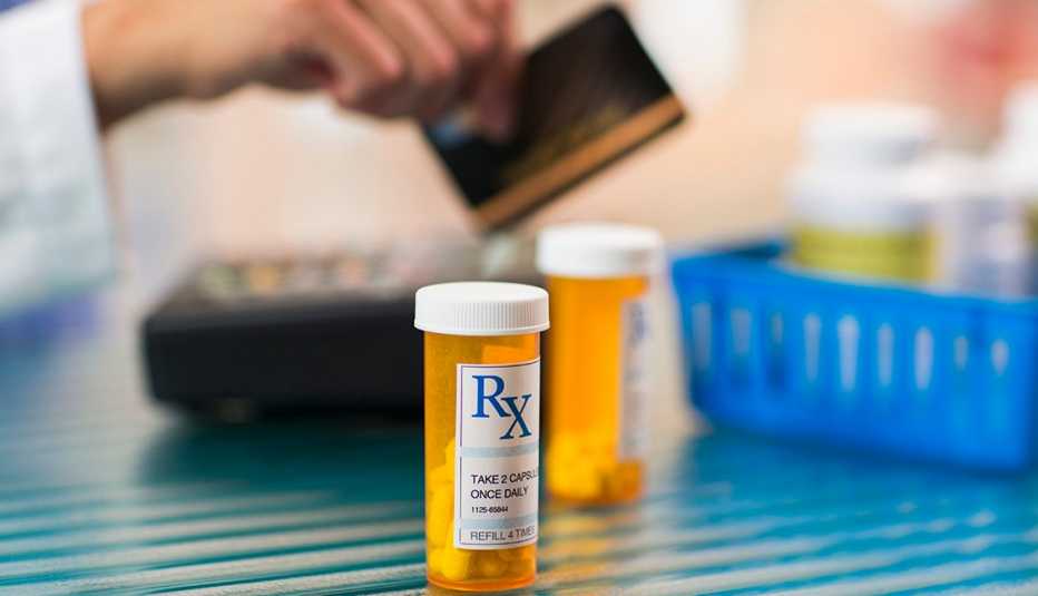 Frasco de medicamentos recetados y en el fondo una persona pagando con tarjeta de crédito