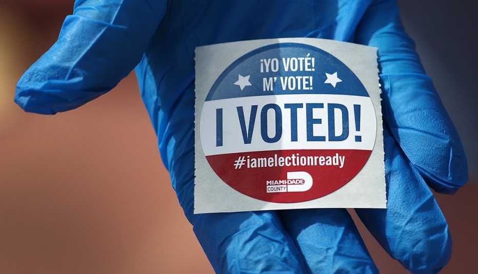 Mano con guante azul sostiene una calcomanía que dice, yo voté