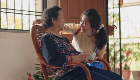 Una mujer mayor sentada tomando té y una mujer joven llega y la saluda Una mujer mayor sentada tomando té y una mujer joven llega y la saluda