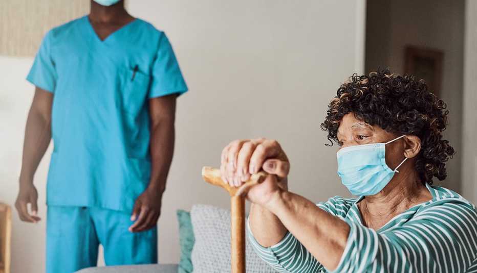 Una mujer residente de un hogar de ancianos sosteniendo un bastón y usando una mascarilla con un trabajador de un hogar de ancianos en el fondo.