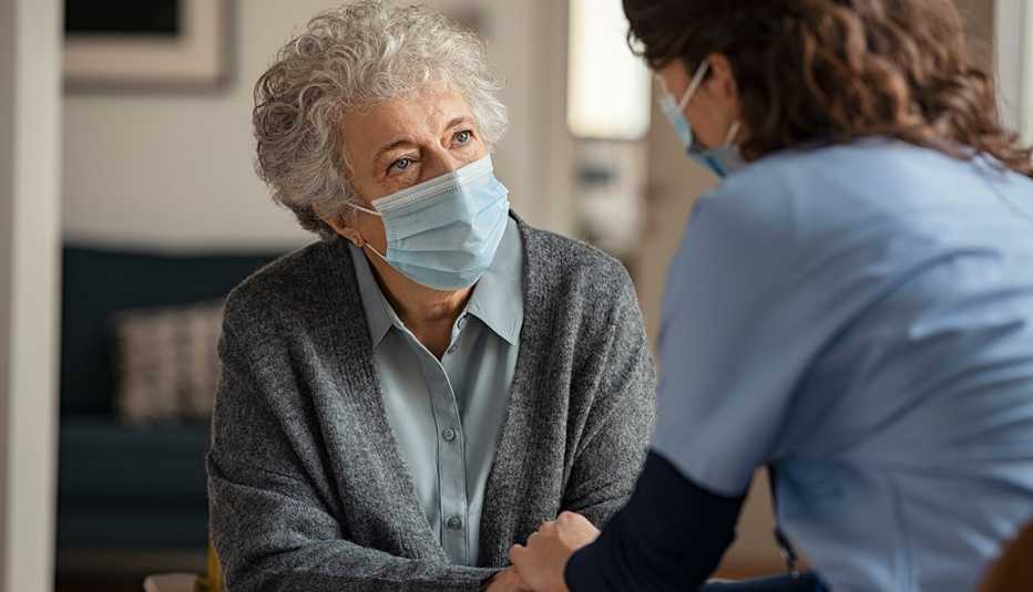 Elderly woman talking with a doctor while holding hands at home and wearing face protective mask. Worried senior woman talking to her general pratictioner visiting her at home during virus epidemic. Doctor explaining about precautionary measures during virus pandemic to old lady and takes care of her.