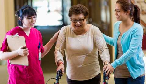 Mujer mayor hispana camina con la ayuda de un caminador y su fisioterapeuta en un hogar de ancianos. La nieta de la mujer también está presente.