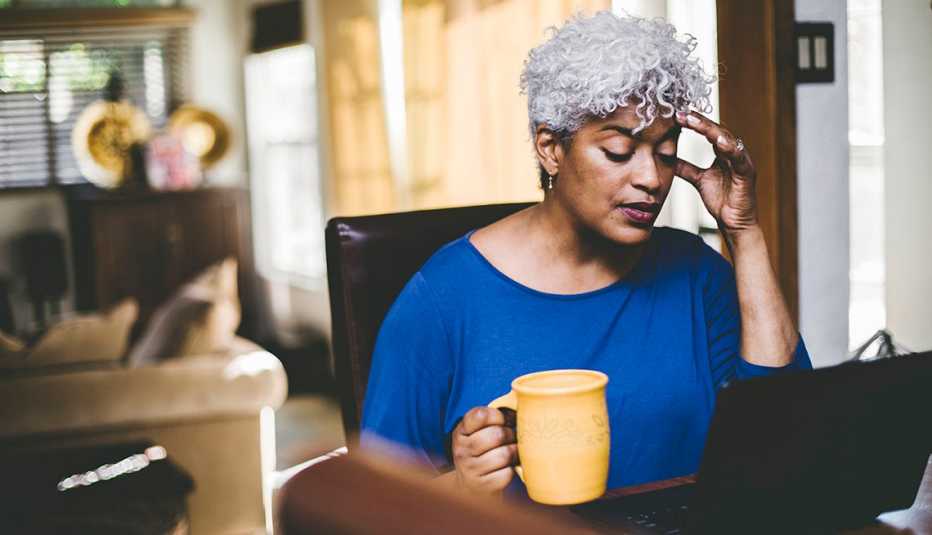 Una mujer trabaja en su computadora, en una mano sostiene una taza y la otra la lleva a su cabeza