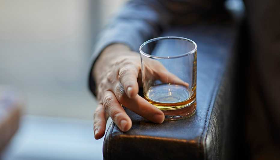 Mano de un hombre en el descanso de una butaca sostiene un vaso con licor Mano de un hombre en el descanso de una butaca sostiene un vaso con licor