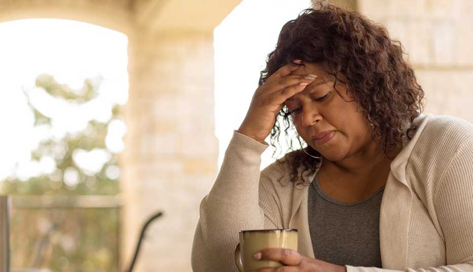 Una mujer luce preocupada, con una mano en la frente mientras en la otra sostiene una taza