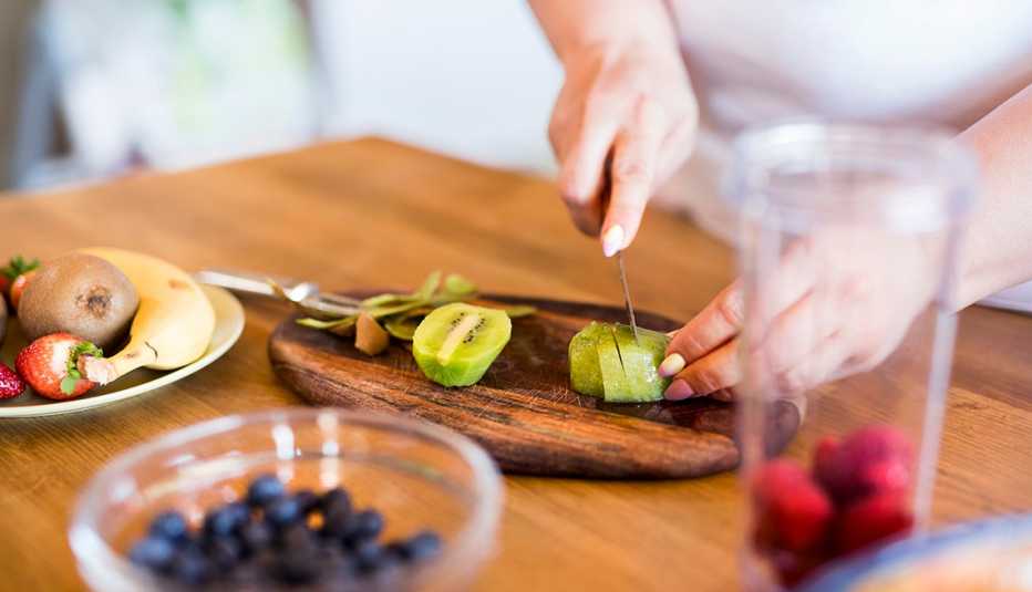 Una mujer corta un kiwi, también hay otros alimentos sobre la mesa