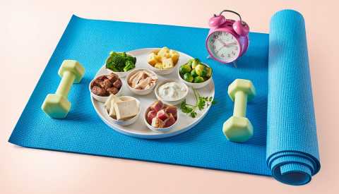 Una estera de yoga y sobre ella hay un reloj, varios alimentos nutritivos y un  par de mancuernas Una estera de yoga y sobre ella hay un reloj, varios alimentos nutritivos y un  par de mancuernas