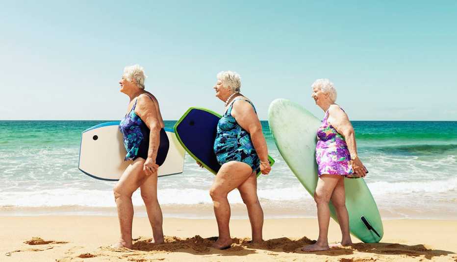 Tres mujeres mayores caminan por una playa cada una con una tabla para practicar el surf Tres mujeres mayores caminan por una playa cada una con una tabla para practicar el surf