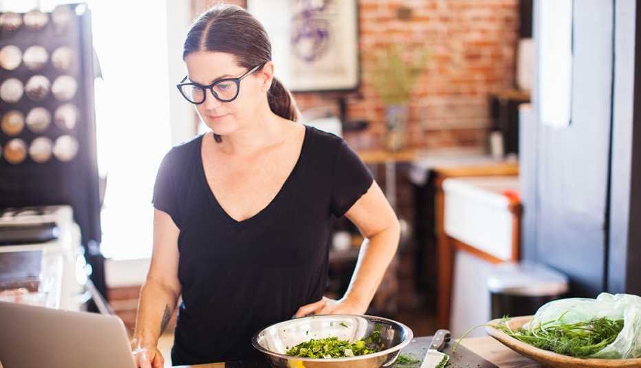 Una mujer mira una computadora mientras prepara una comida en su cocina