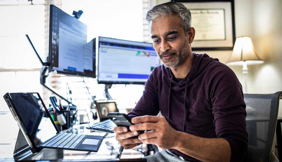 Hombre trabajando desde su casa con dos monitores, una computadora portátil y mirando un teléfono móvil.