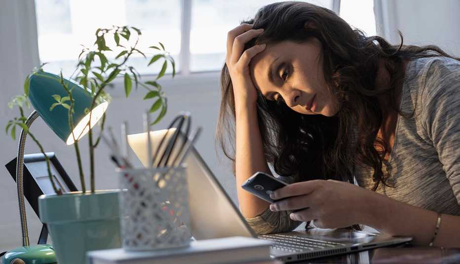 Mujer mira su teléfono móvil mientras apoya su mano en un escritorio y se sostiene la cabeza.