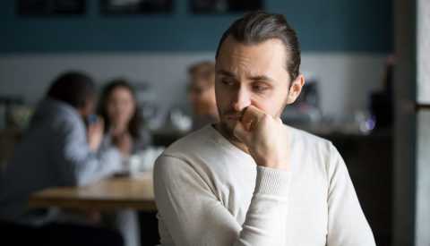 Hombre sentado en una cafetería mientras se lleva la mano a la boca y mantiene su mirada hacia abajo. 