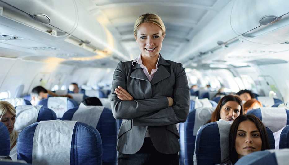 Una asistente de vuelo sonrie de pie en el pasillo de un avión lleno de pasajeros