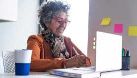 A woman of approximately 60 years of age with gray hair dressed in formal clothes is sitting at the desk in her apartment using her laptop to work remotely, given the covid-19 virus on her desk, we look at her pencils, markers, and notepad