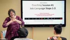Partner with Us Photo Female instructor with brown curly hair wearing purple shirt with a TV screen in classroom