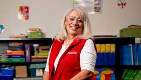 AARP Foundation Experience Corps volunteer tutor Nina Sanchez, older senior woman smiling and standing inside classroom wearing a red vest  