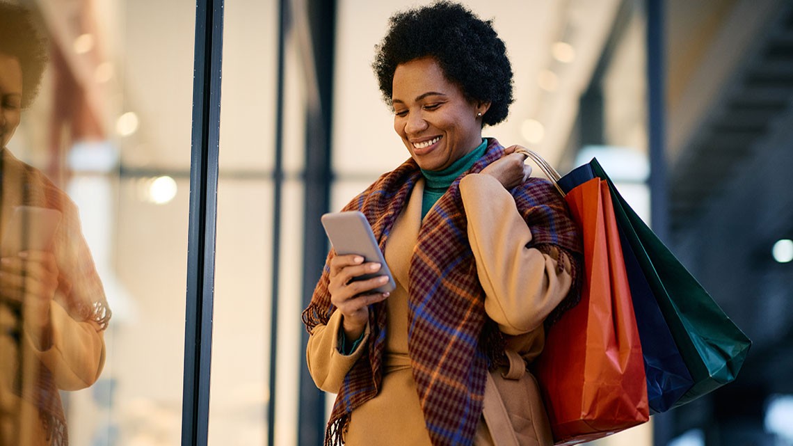 Woman wearing winter coat and scarf, turtleneck, outside shop window holding shopping bags, looking and smiling at cellphone