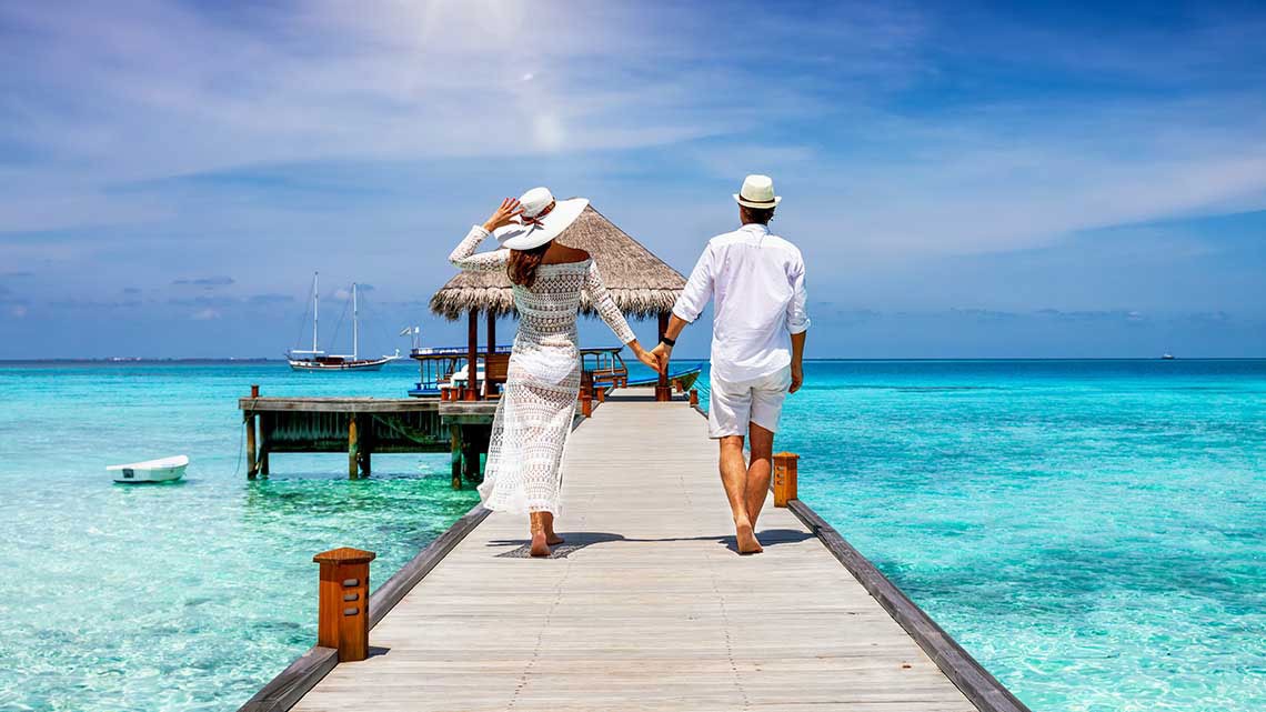 A happy couple in white summer clothing on vacation walks along a wooden pier over tropical, turquoise ocean in the Maldives, Indian Ocean