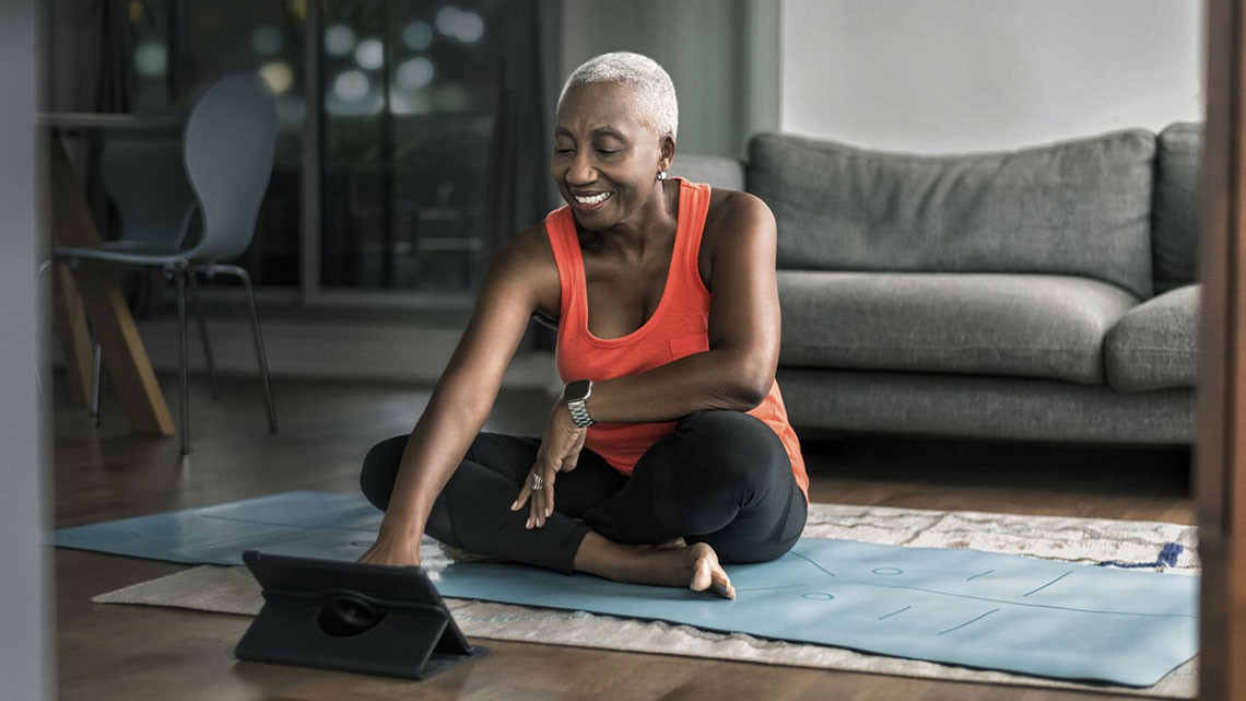 african american older woman sitting with crossed legs on yoga mat with tablet on floor