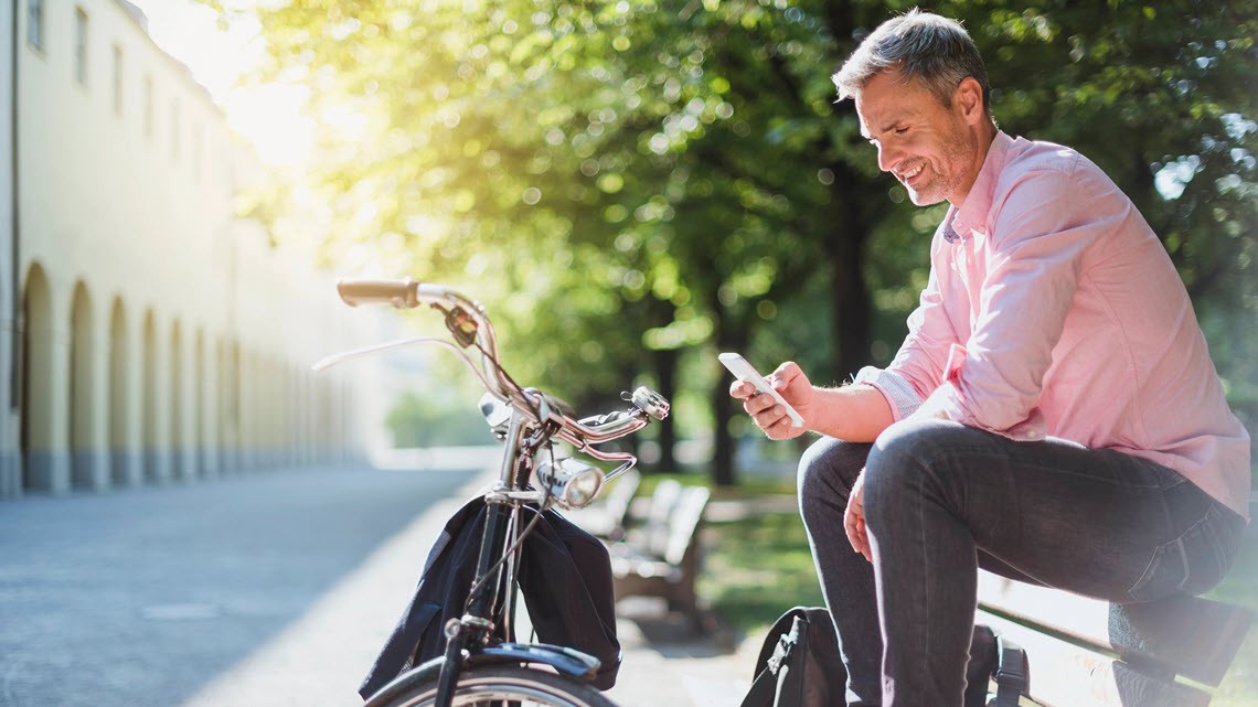 man sitting on bench looking at phone with bicycle standing in front of him