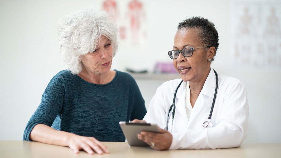older woman with white hair talking to older female doctor in white coat using a tablet in doctor office setting