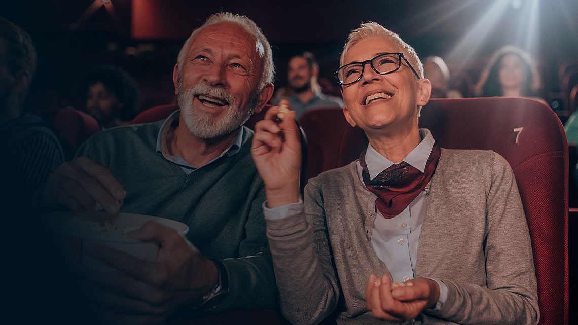 older couple, man and woman, sitting next to each other in a movie theater, eating popcorn, smiling