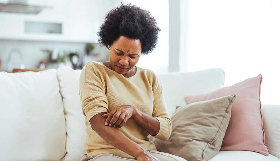 A woman is seated on a cozy sofa, scratching her arm, indicating skin irritation or discomfort. The setting is a bright and casual living room, conveying a sense of home and everyday life.