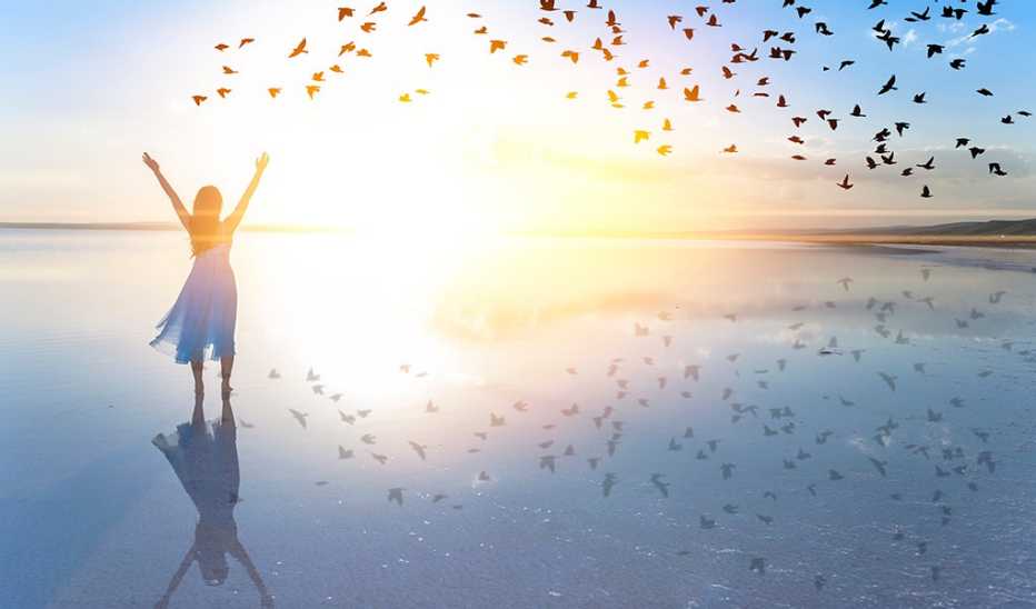 A woman with her arms raised standing by a lake at sunset
