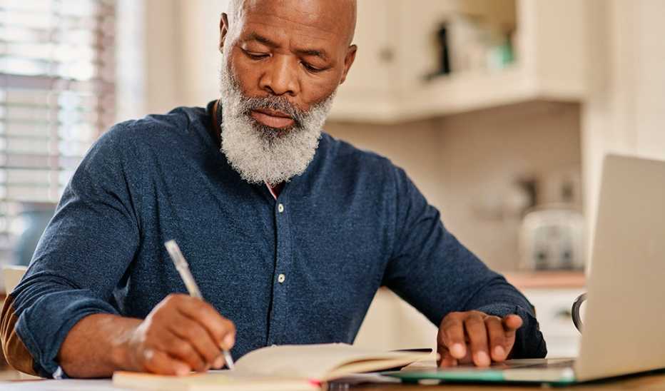 A man writing in a journal with a laptop in front of him