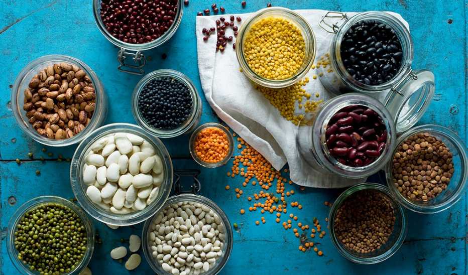 An overhead view of various beans and lentils in jars on blue background