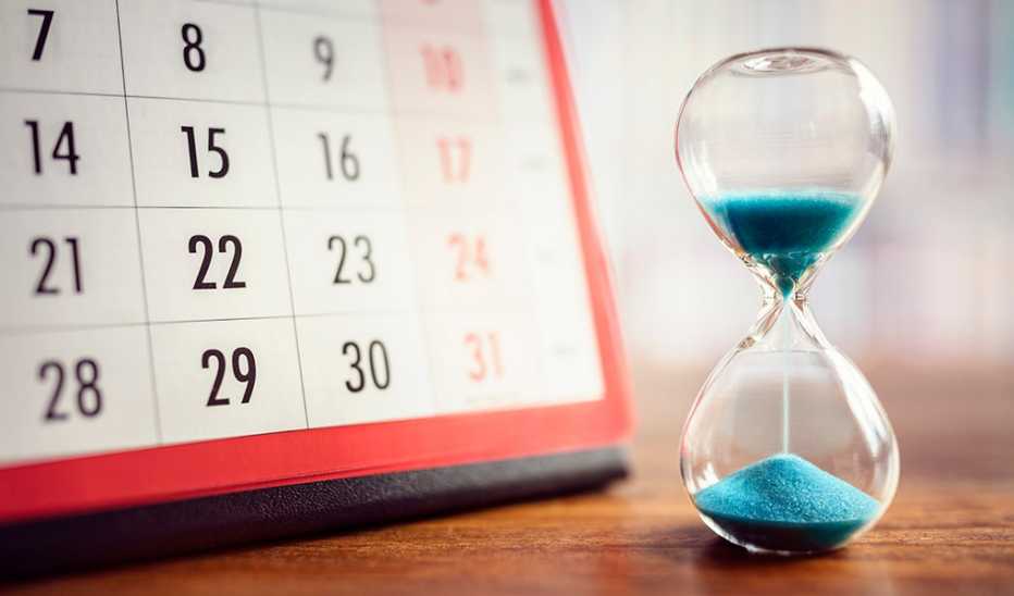 An hourglass with sand pouring down and a calendar beside it on a wooden table