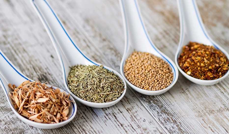 A close-up view of spices and herbs in porcelain spoons over a wooden background
